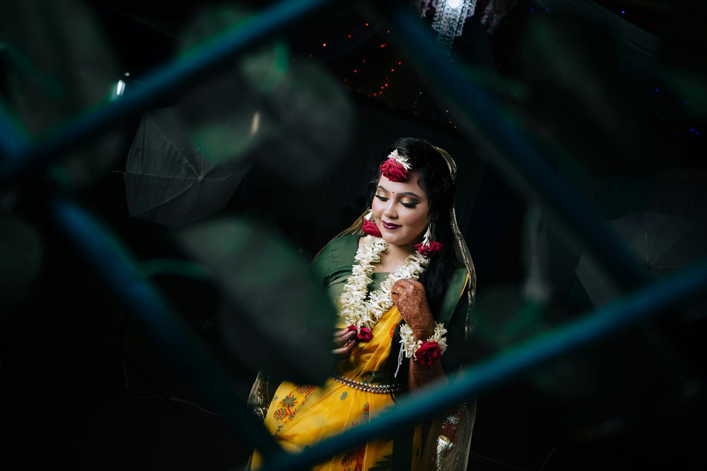 A bride in a yellow sari and flower garlands, smiling down, framed by dark leaves at night