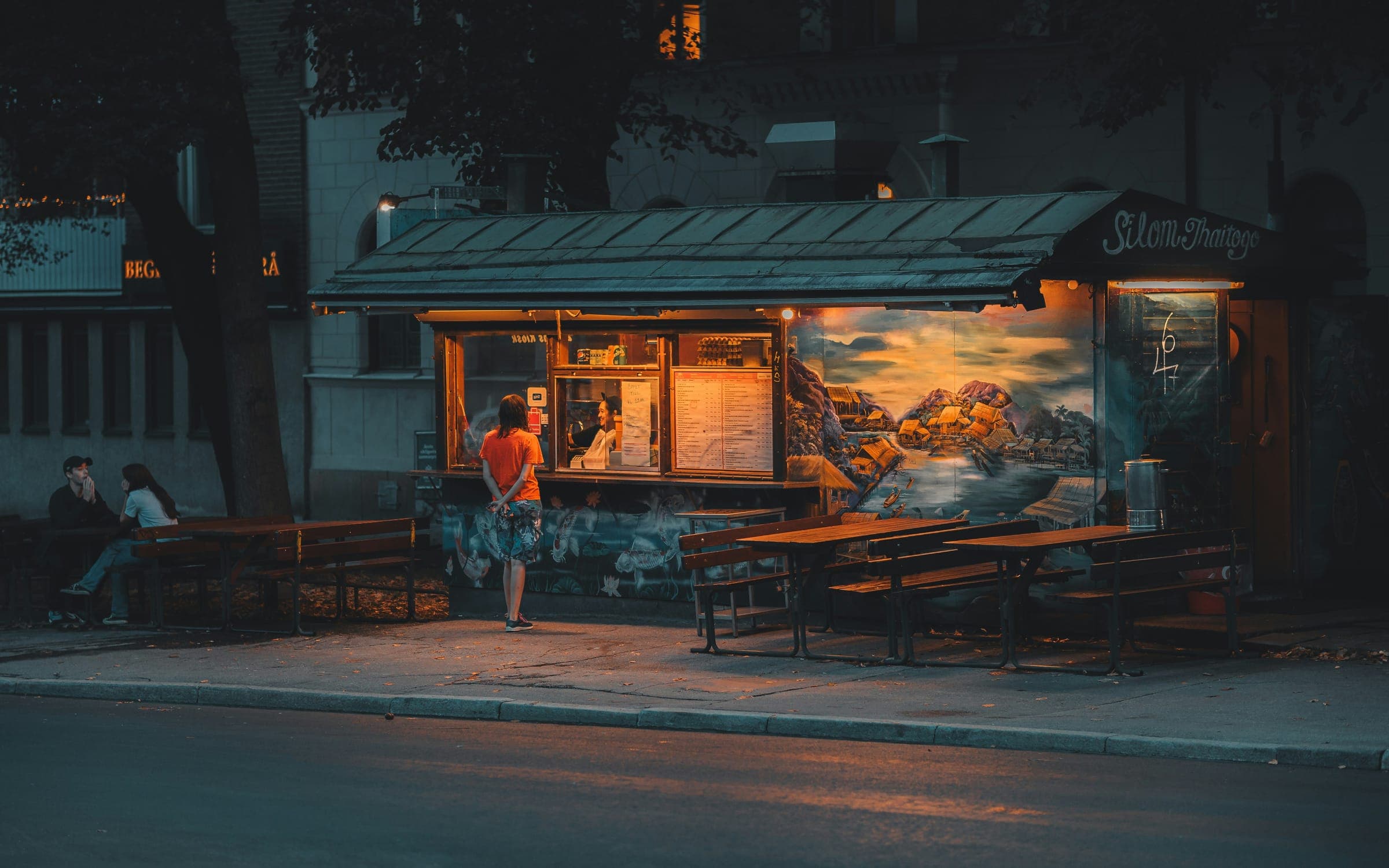 A lit food kiosk on a quiet street at dusk, one customer at the window