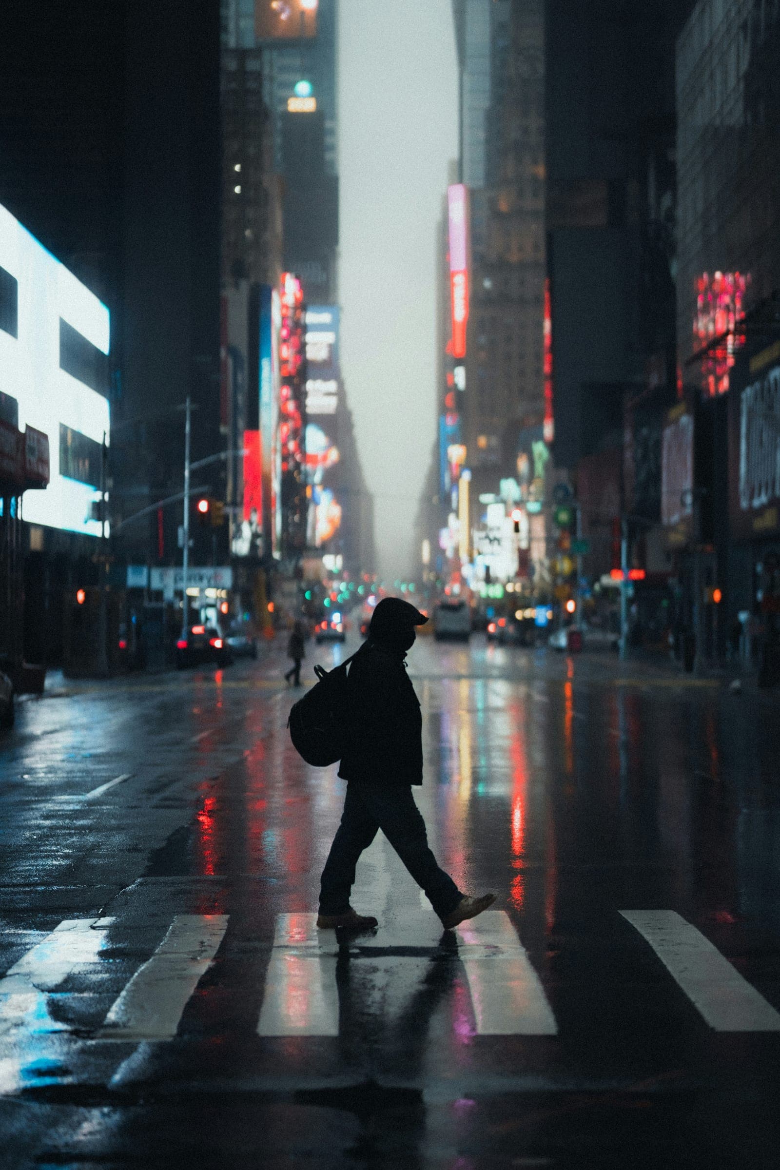 A figure with a backpack crosses a wet city street at night under neon signs