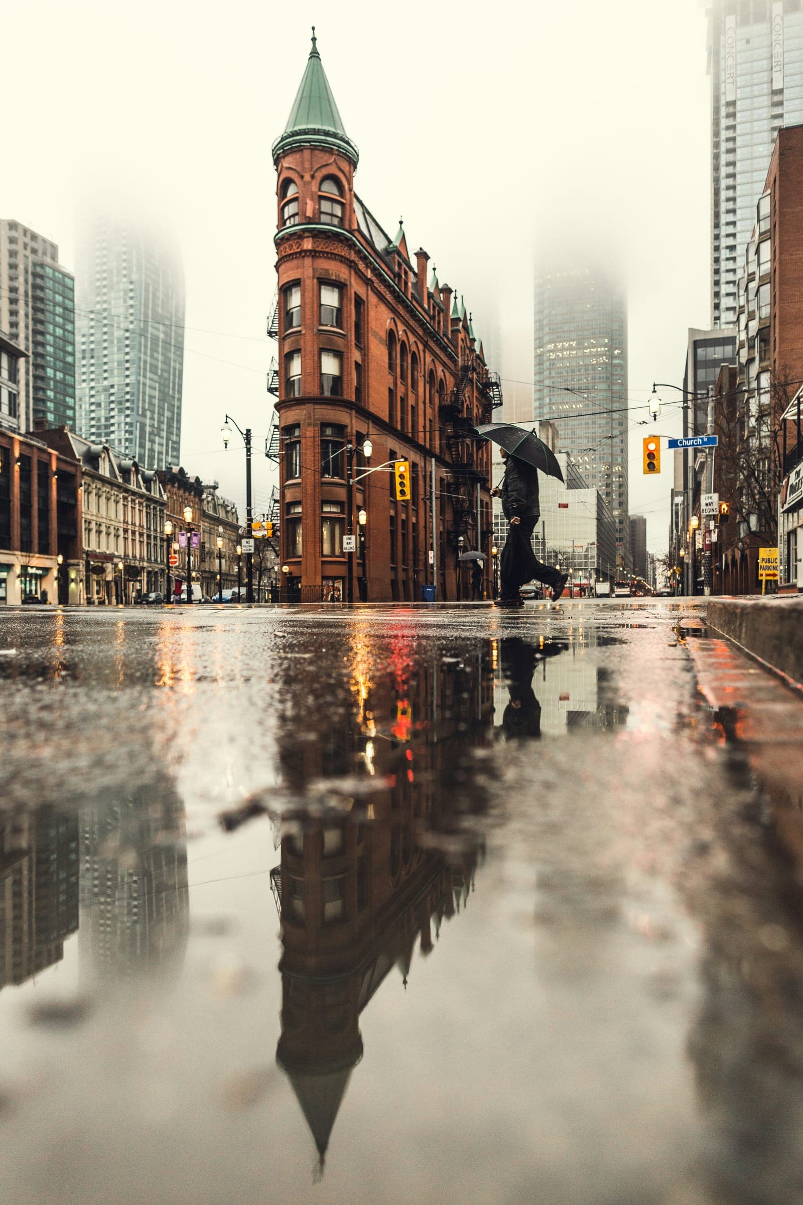 A red-brick corner building and its reflection in a rain puddle, towers fading into fog