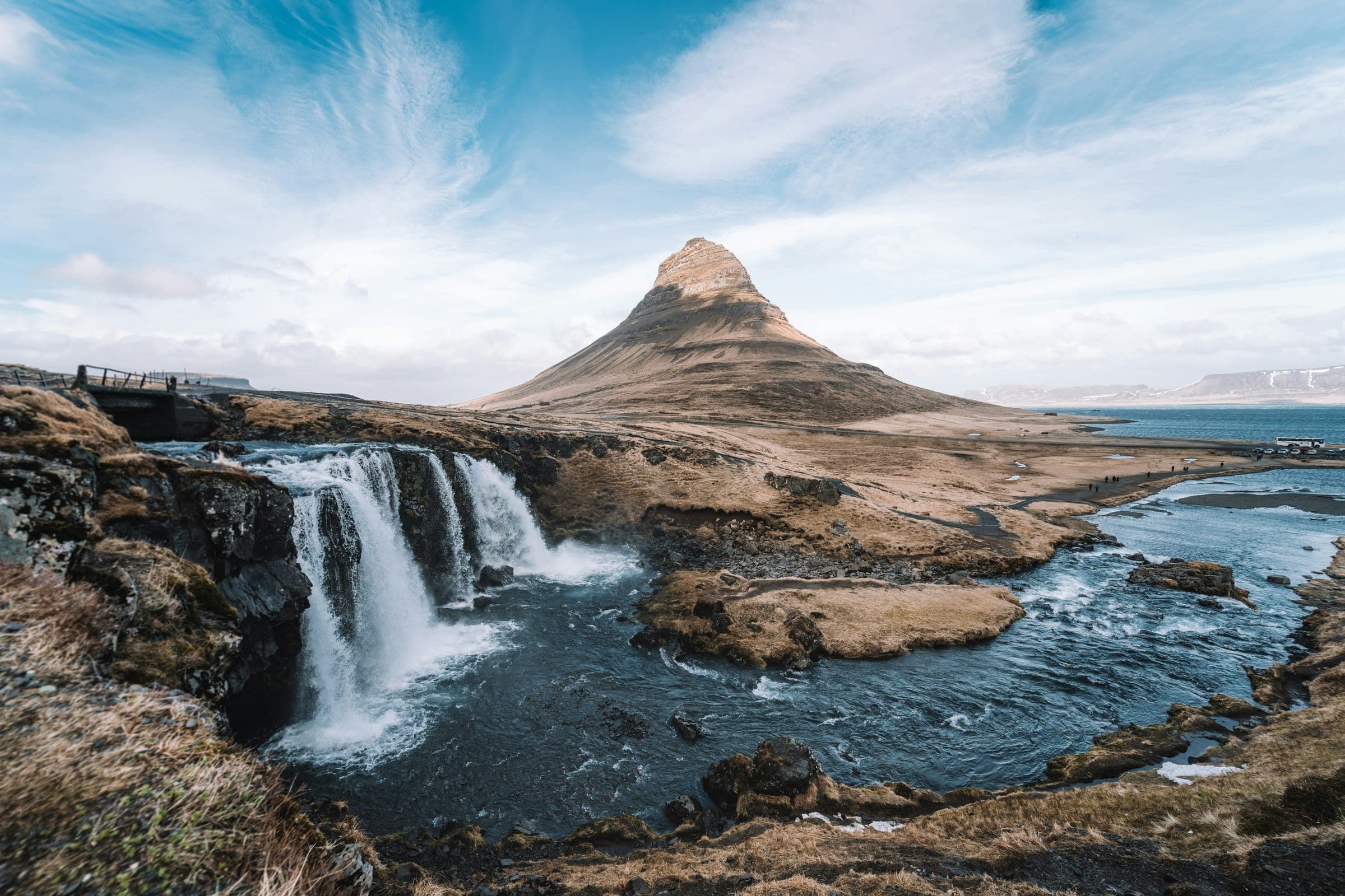 A waterfall in front of a cone-shaped mountain by the sea, under a wide blue sky