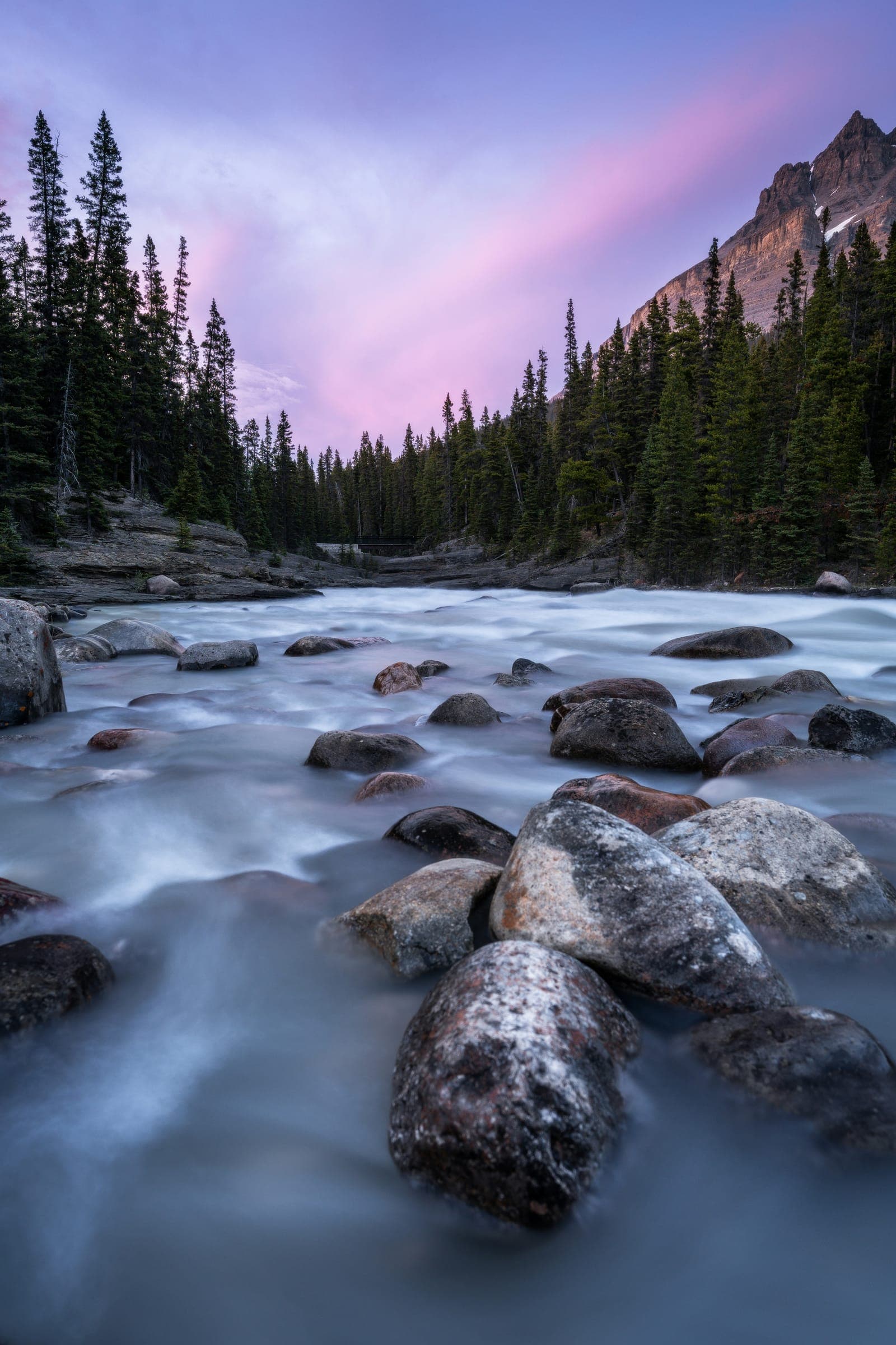 Long exposure of a river smoothing around boulders, pines and a pink sky beyond