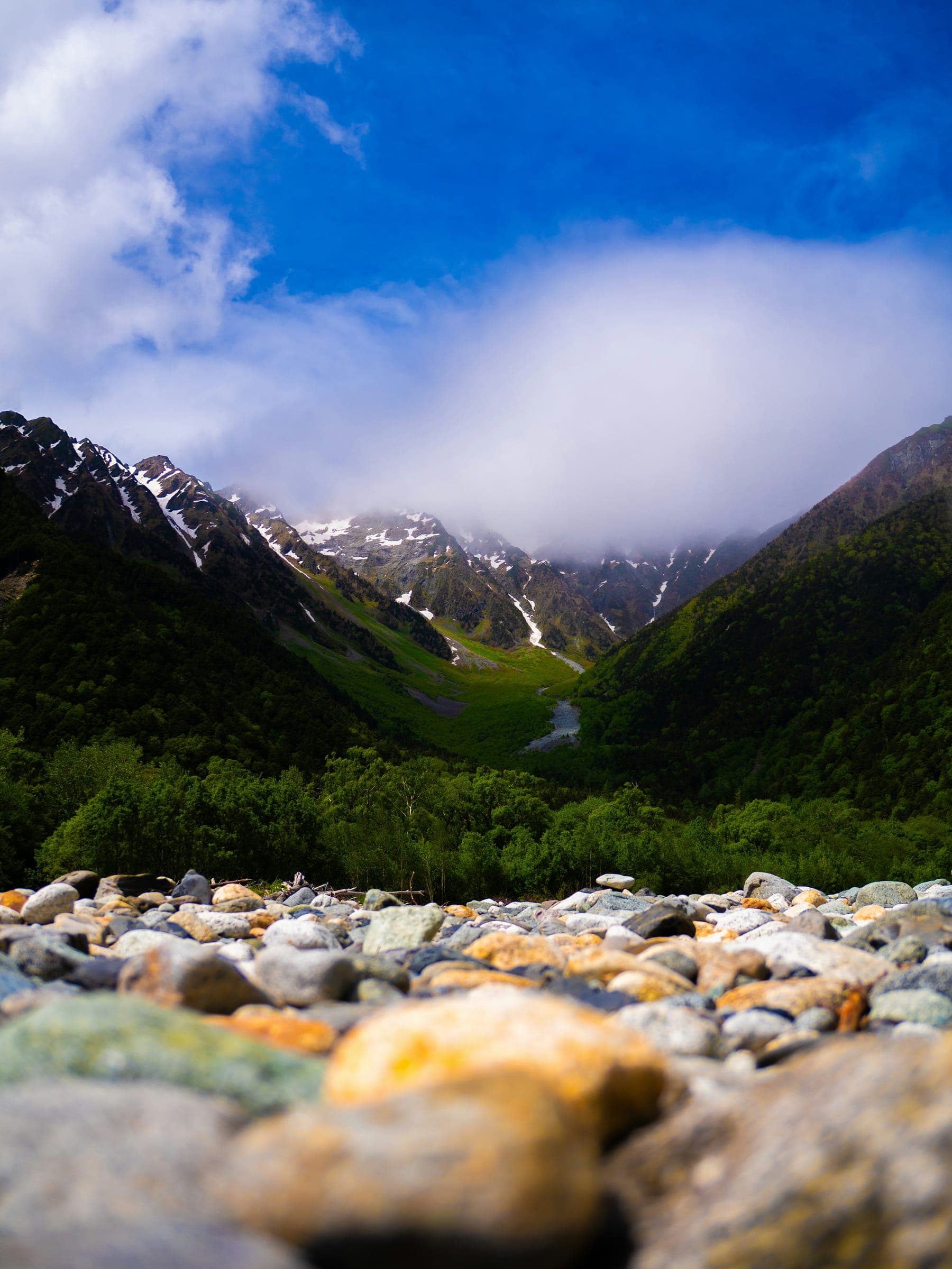 A green mountain valley under a blue sky, smooth pebbles in the foreground