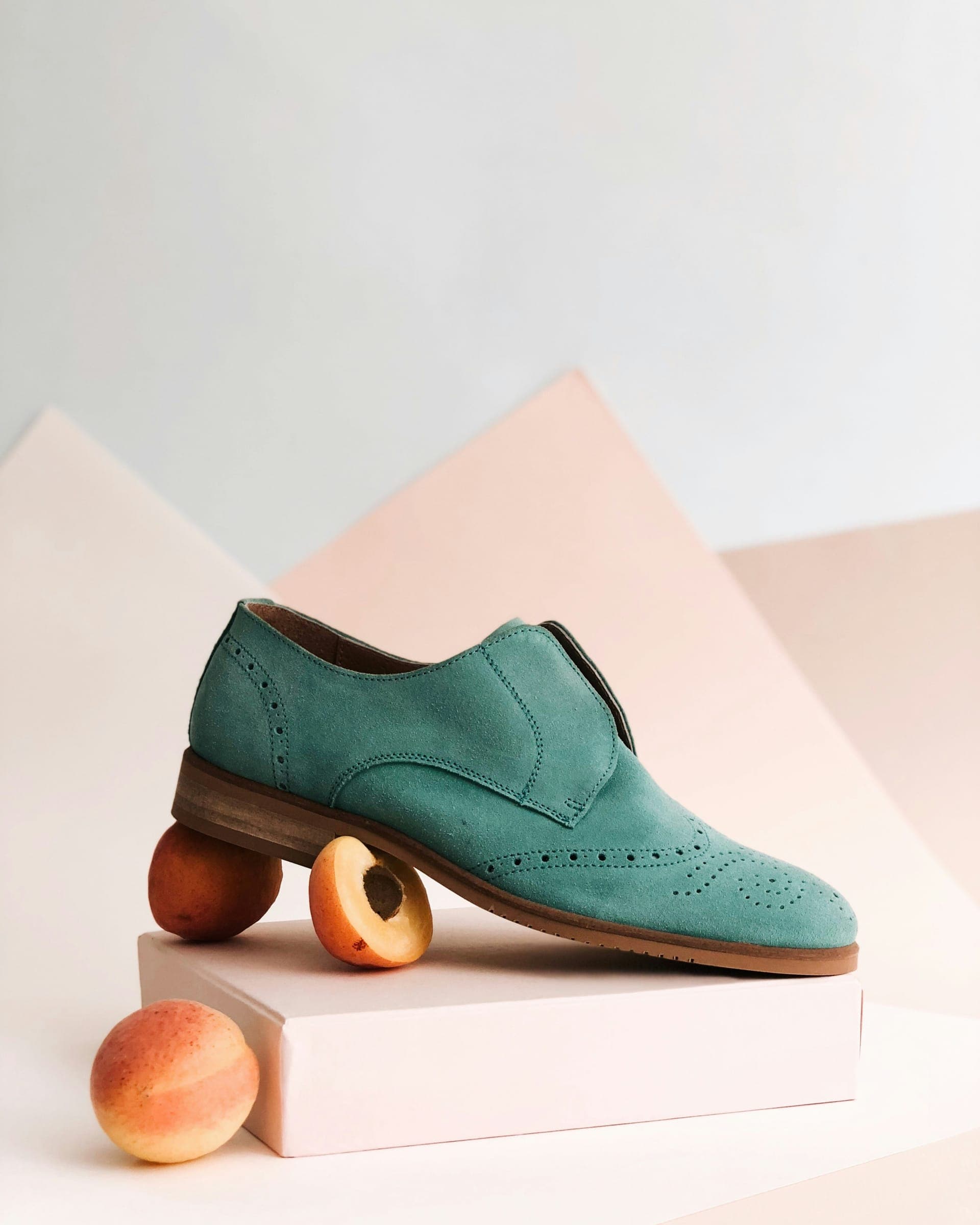 A teal suede brogue balanced on peaches on a pale pink plinth