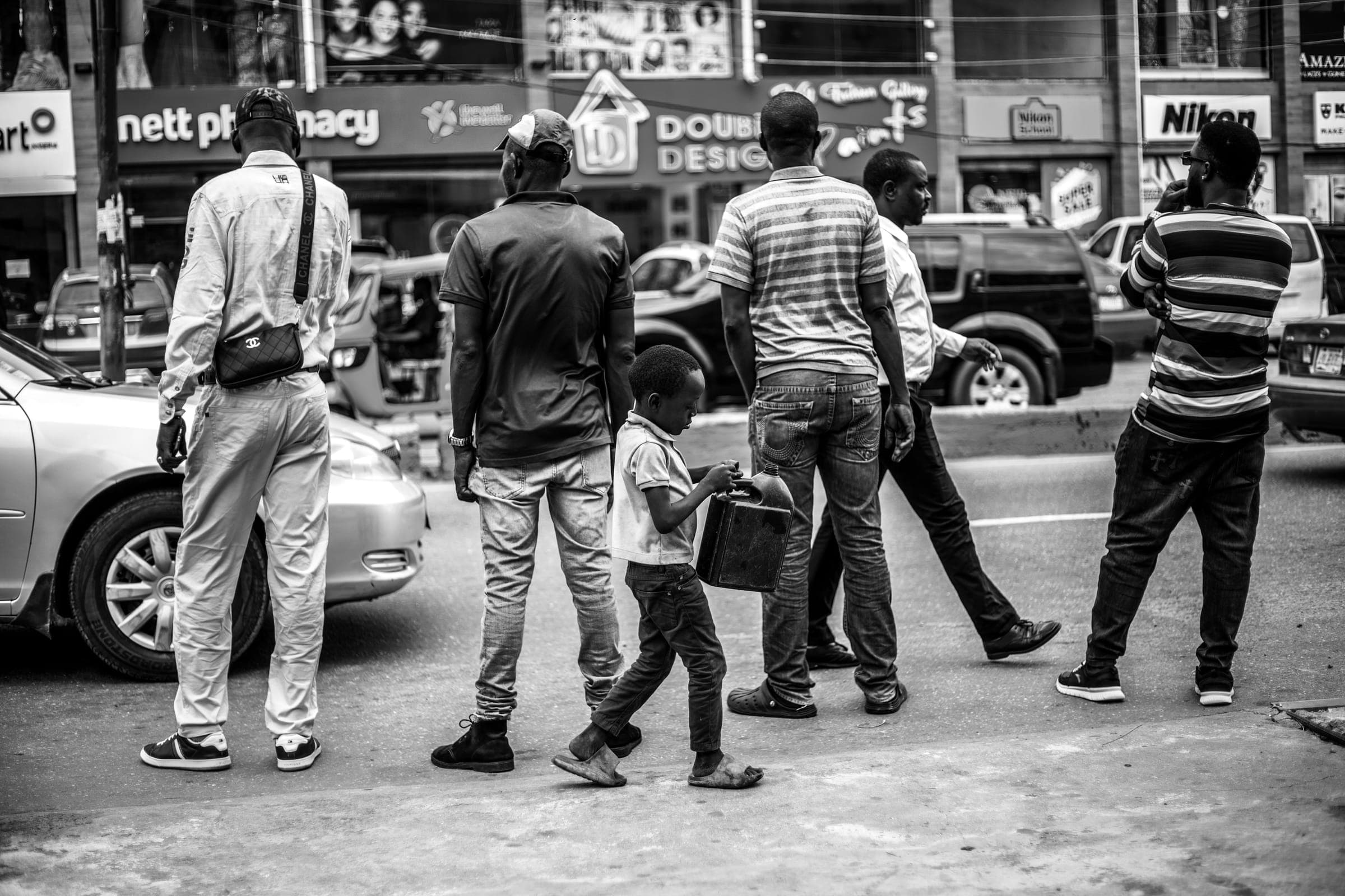 Black and white: men and a boy cross a busy street, shop signs behind them