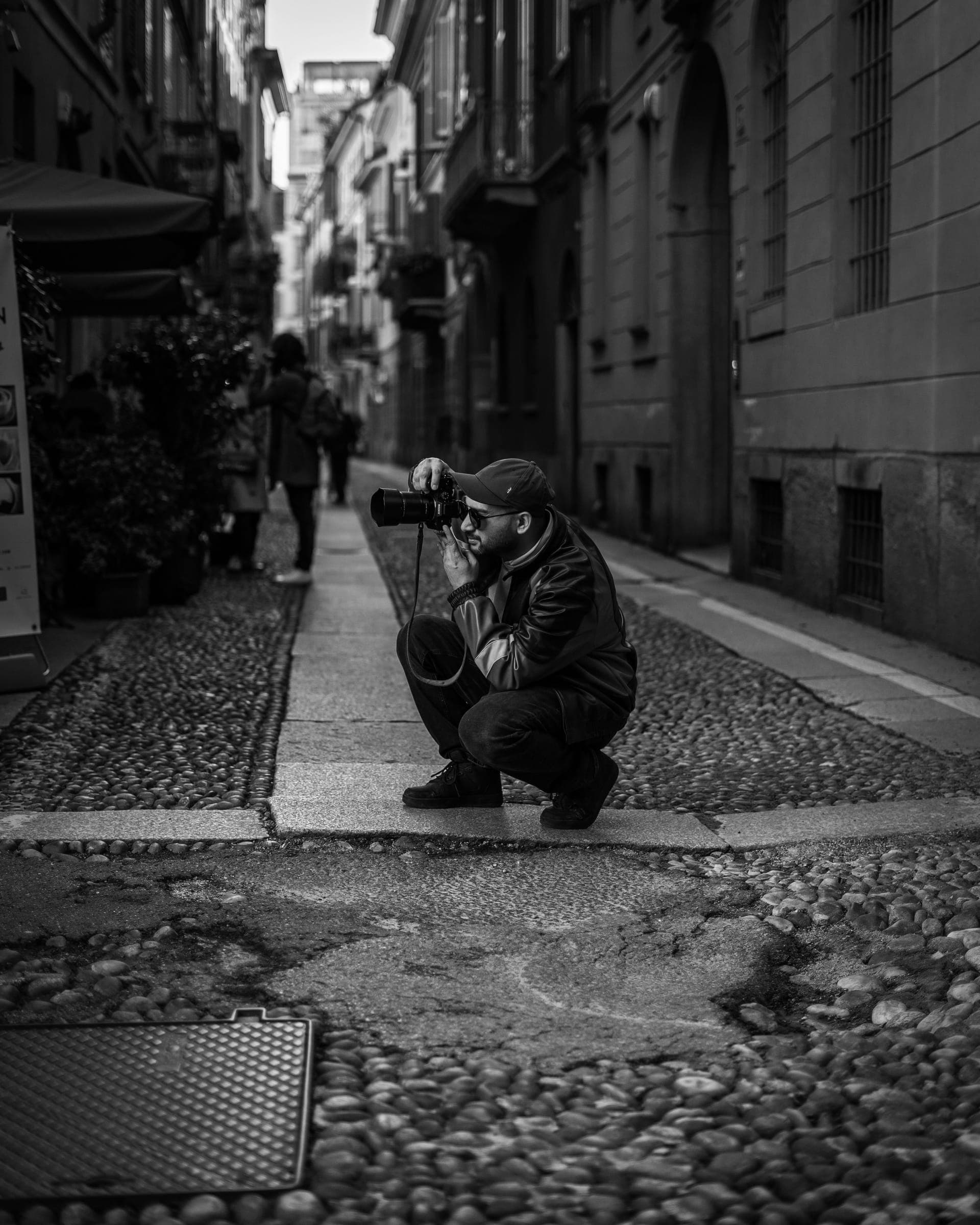 Black and white: a photographer crouches on a cobbled lane, camera raised