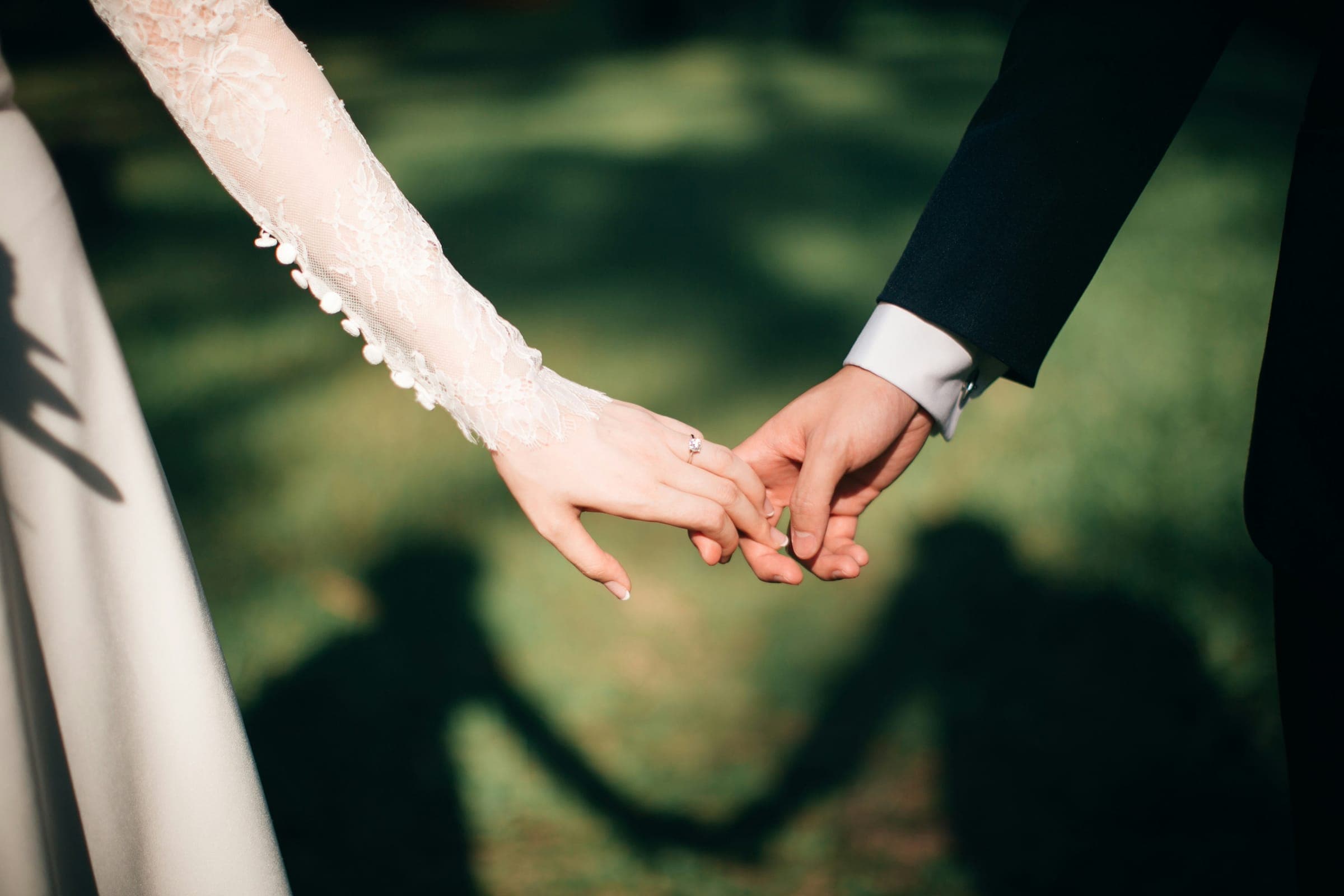 A bride and groom reach for each other's hands, their shadows stretched across the lawn