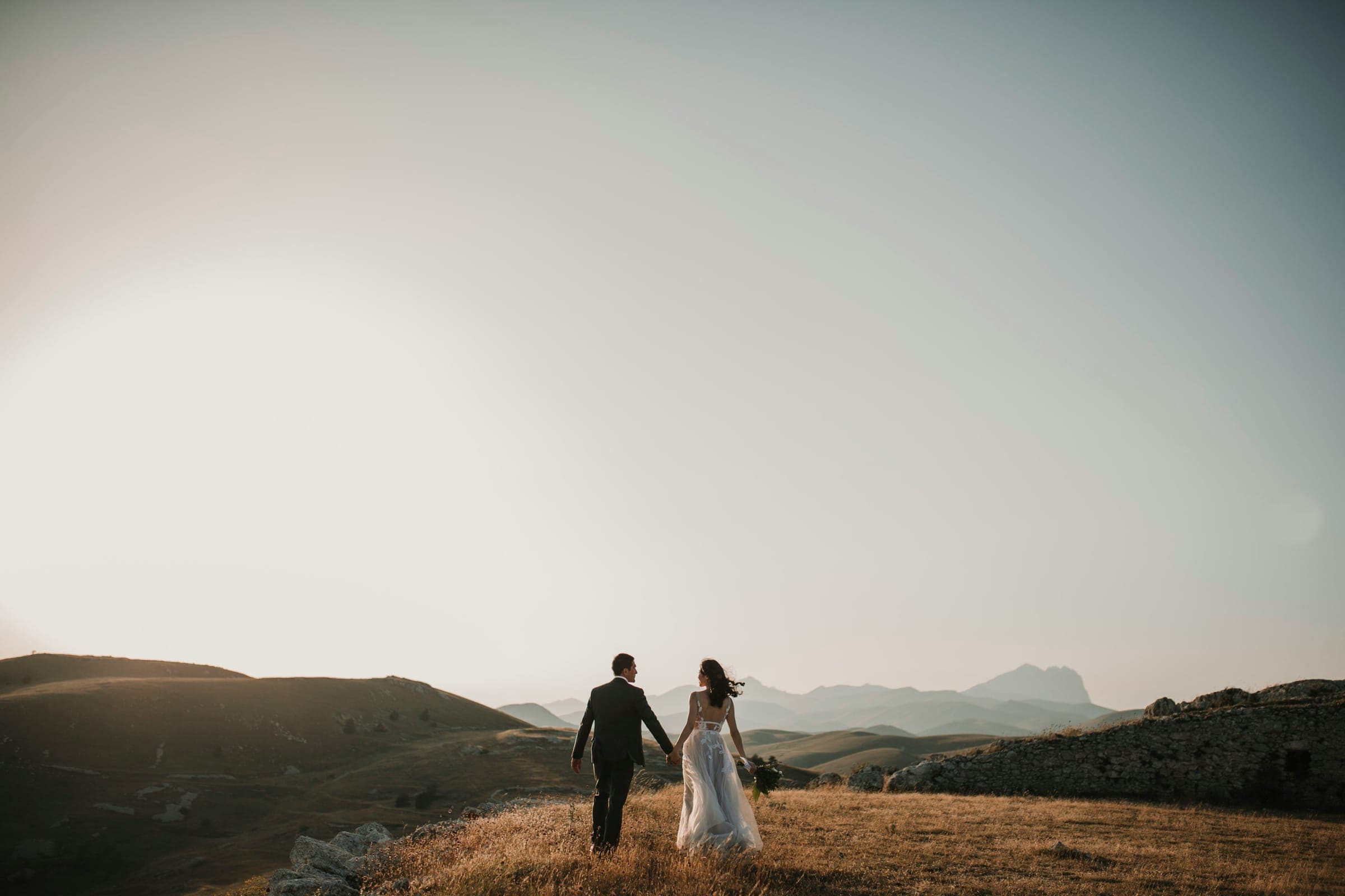 A bride and groom walk hand in hand across a hilltop at golden hour, mountains behind them